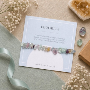 May include: A bracelet made of small, polished, multi-coloured fluorite stone chips attached to a light blue display card. The card features the text FLUORITE, a description of the stone, and the text MOODLIFT MAIL at the bottom in black font.