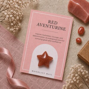 May include: A star-shaped red aventurine stone attached to a rectangular pink card. The card features the text RED AVENTURINE, a description of the stone, and MOODLIFT MAIL at the bottom. The stone has a smooth, polished, reddish-brown surface.