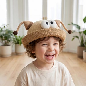 Może przedstawiać: Beżowy słomkowy kapelusz typu bucket hat z dwoma dużymi białymi oczami z kreskówki z czarnymi źrenicami z przodu oraz dwiema długimi, zakrzywionymi antenkami po bokach, z których każda zakończona jest małą brązową kulką, co nadaje mu zabawny wygląd.