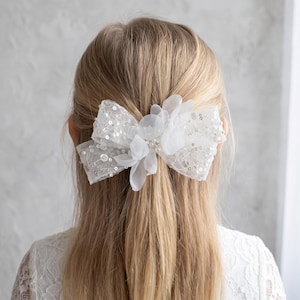 May include: A white hair bow featuring a sheer lace fabric base adorned with scattered silver sequins and small white fabric petals. The centre of the bow is embellished with a cluster of small white faux pearls, creating a textured accessory.