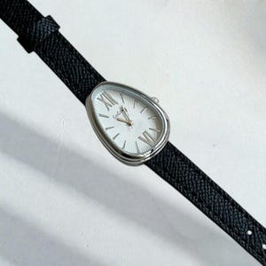 Puede incluir: Un reloj plateado con esfera blanca y correa de cuero negro. La esfera del reloj tiene números romanos y la marca "GoGo". La correa es texturizada y el reloj tiene forma ovalada.