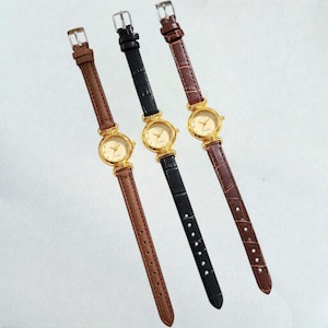 Puede incluir: Tres relojes de pulsera dorados con correas de cuero en marrón y negro. Cada reloj tiene una esfera redonda con manecillas doradas y un bisel dorado. Las correas tienen un patrón texturizado similar al de un cocodrilo.