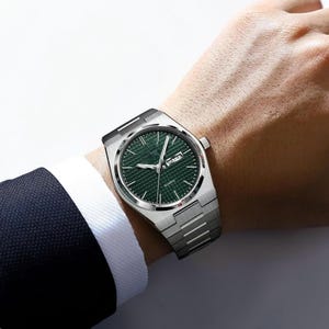 May include: A silver-toned wristwatch with a green face and a date display. The watch has a textured dial and a stainless steel band. The watch is being worn on a wrist, with a dark suit jacket visible in the background.