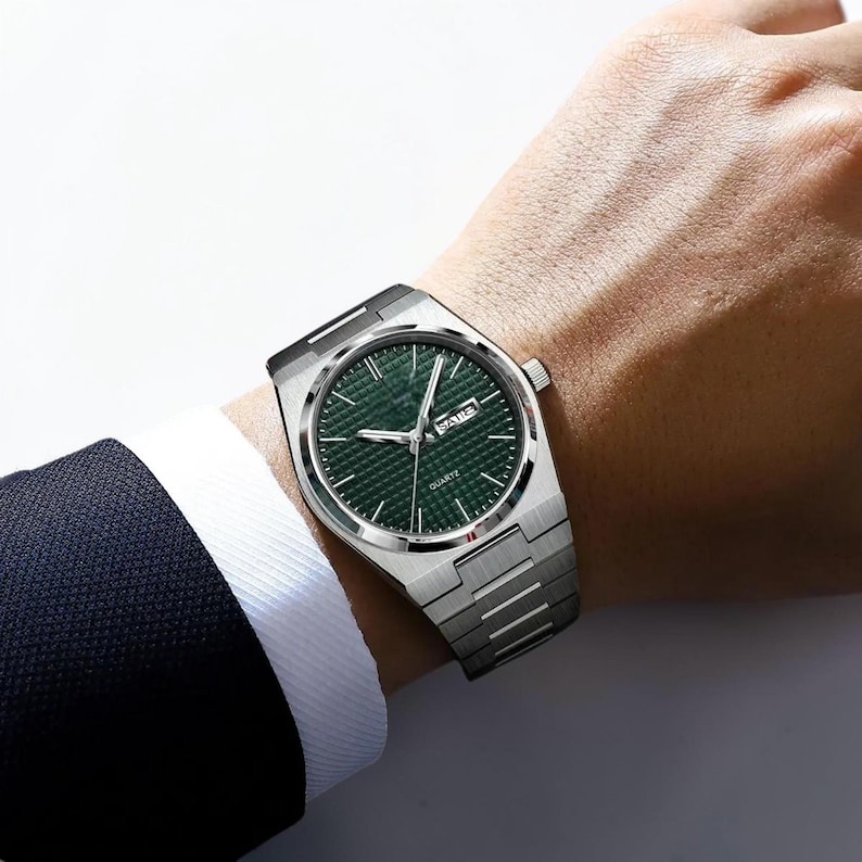 May include: A silver-toned wristwatch with a green face and a date display. The watch has a textured dial and a stainless steel band. The watch is being worn on a wrist, with a dark suit jacket visible in the background.