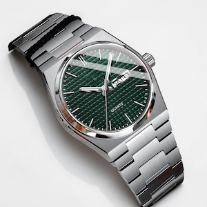 May include: A silver-toned wristwatch with a green textured dial. The watch features a rectangular face with the day of the week displayed. The watchband is made of silver-toned metal links. The word "QUARTZ" is printed on the watch face.