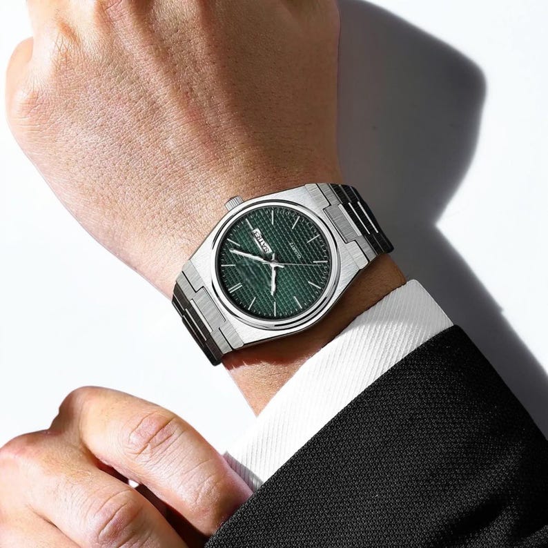 May include: A silver wristwatch with a green textured dial is displayed on a wrist. The watch has a rectangular case and a silver band. The watch is being worn with a black suit and white shirt.