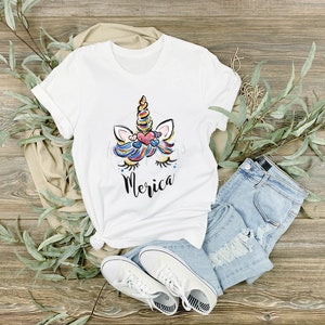 May include: White t-shirt featuring a colourful unicorn graphic and the word "Merica" printed on the front. The shirt is laid out on a wooden surface with blue jeans, white trainers, and green leaves.