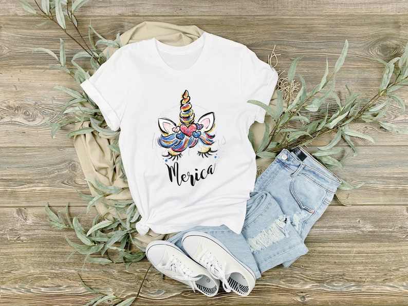 May include: White t-shirt featuring a colourful unicorn graphic and the word "Merica" printed on the front. The shirt is laid out on a wooden surface with blue jeans, white trainers, and green leaves.