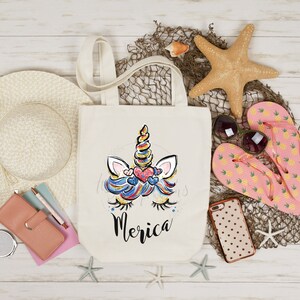 May include: A white canvas tote bag featuring a colourful unicorn face and the word "Merica" printed on it. The bag is surrounded by beach-themed items such as a straw hat, flip flops, a starfish, and a seashell.