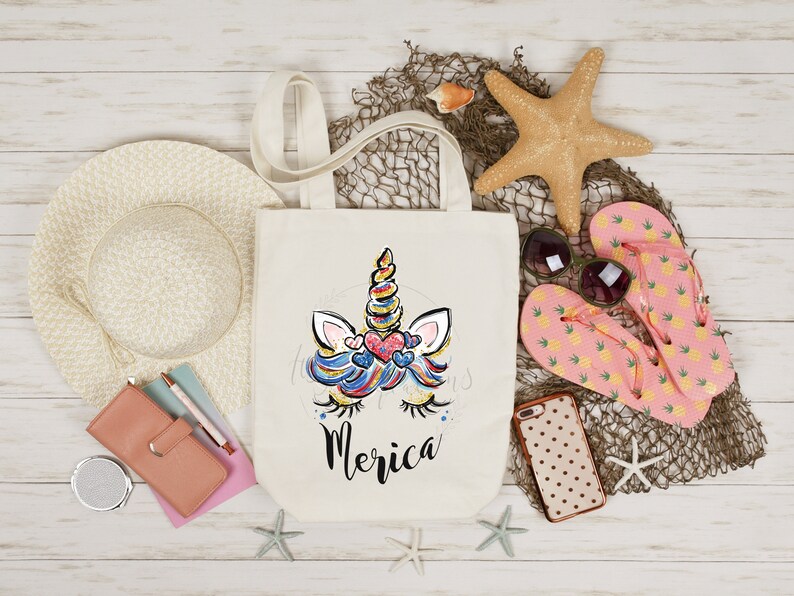 May include: A white canvas tote bag featuring a colourful unicorn face and the word "Merica" printed on it. The bag is surrounded by beach-themed items such as a straw hat, flip flops, a starfish, and a seashell.