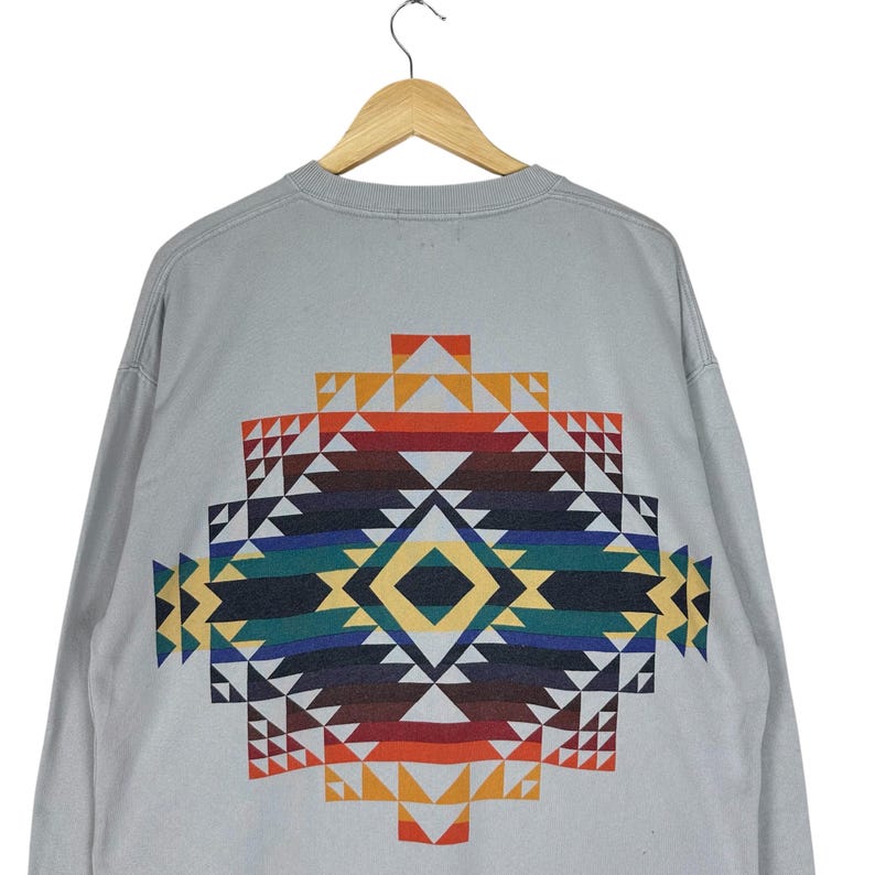 Puede incluir: Sudadera de manga larga gris claro con un colorido estampado geom&eacute;trico en la espalda. El patr&oacute;n presenta tri&aacute;ngulos en tonos rojos, naranjas, amarillos, verdes, azules y marrones, creando un dise&ntilde;o llamativo.