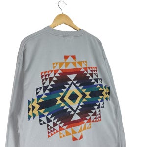 Puede incluir: Una sudadera gris claro con un colorido dise&ntilde;o geom&eacute;trico en la espalda. El dise&ntilde;o presenta una forma de diamante con tri&aacute;ngulos en tonos rojos, naranjas, amarillos, verdes, azules y marrones. La sudadera est&aacute; colgada en una percha de madera.