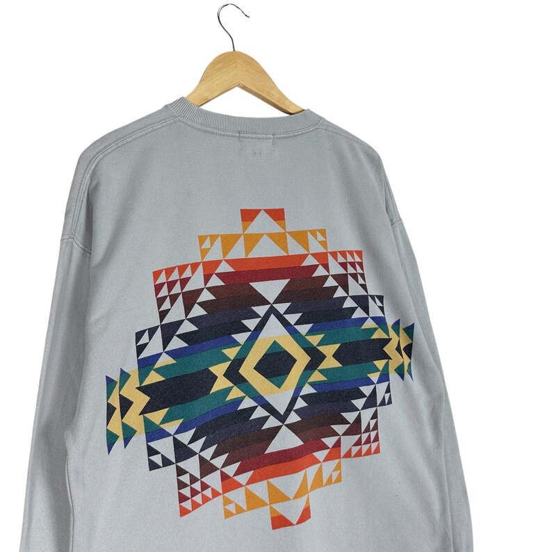 Puede incluir: Una sudadera gris claro con un colorido dise&ntilde;o geom&eacute;trico en la espalda. El dise&ntilde;o presenta una forma de diamante con tri&aacute;ngulos en tonos rojos, naranjas, amarillos, verdes, azules y marrones. La sudadera est&aacute; colgada en una percha de madera.