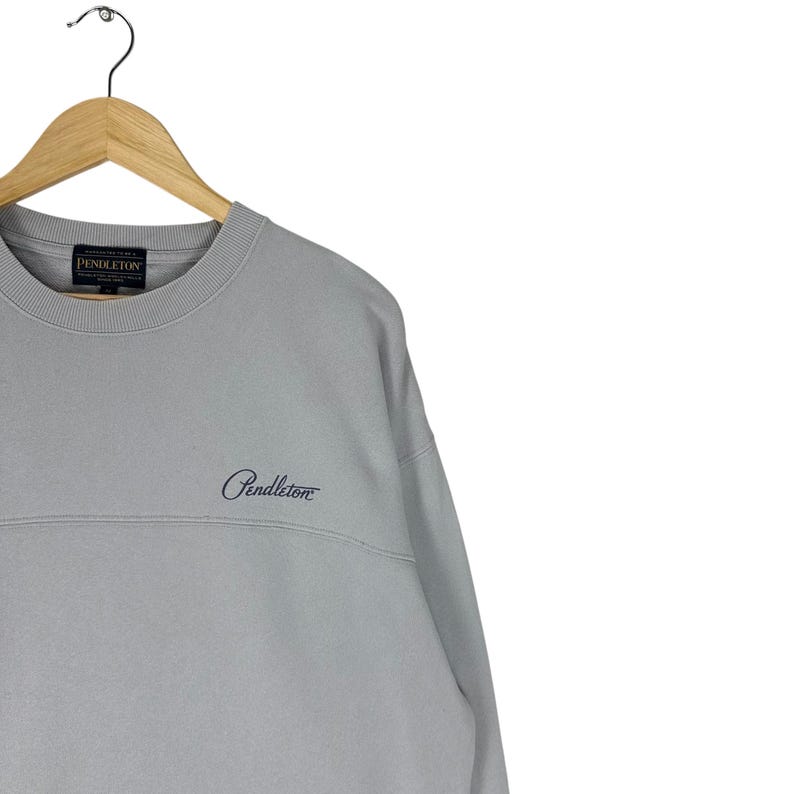 Puede incluir: Una sudadera gris claro con cuello redondo y el nombre de la marca "Pendleton" bordado en el pecho. La sudadera est&aacute; colgada en una percha de madera.