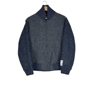 May include: A charcoal gray bomber jacket featuring a textured, woven grid pattern on the front panels, solid dark gray sleeves, a ribbed collar, cuffs, and hem, a front zipper closure, and a small white rectangular brand label on the lower left sleeve.