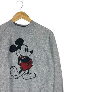 May include: A gray sweatshirt featuring a large graphic of Mickey Mouse. The cartoon character is depicted in black, red, and white. The sweatshirt is hanging on a wooden hanger against a white background.