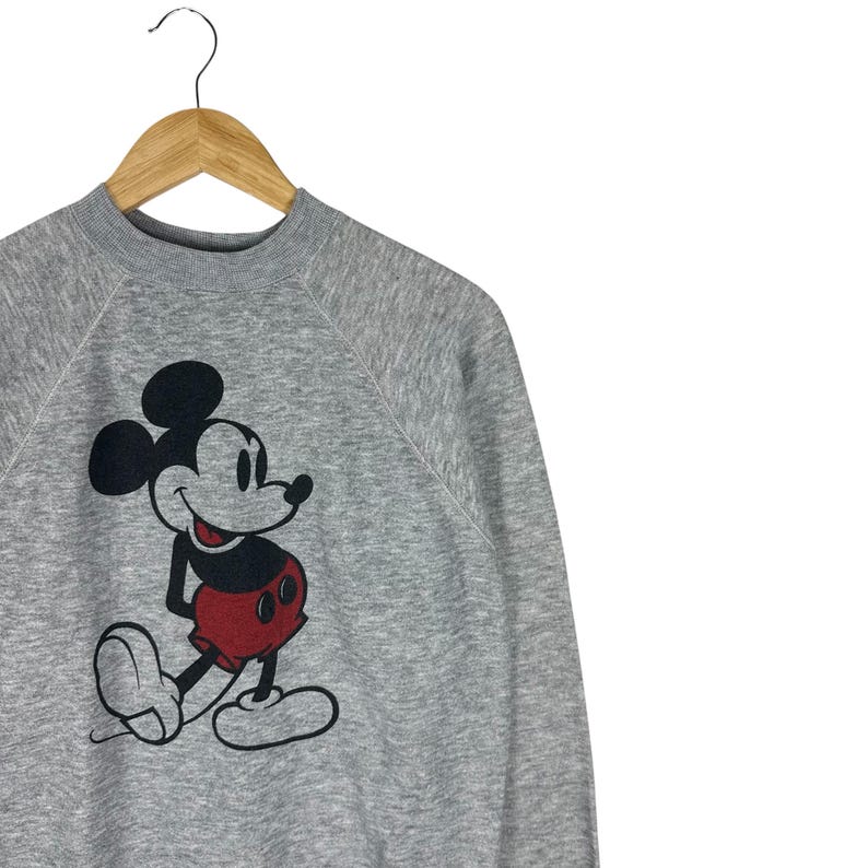 May include: A gray sweatshirt featuring a large graphic of Mickey Mouse. The cartoon character is depicted in black, red, and white. The sweatshirt is hanging on a wooden hanger against a white background.