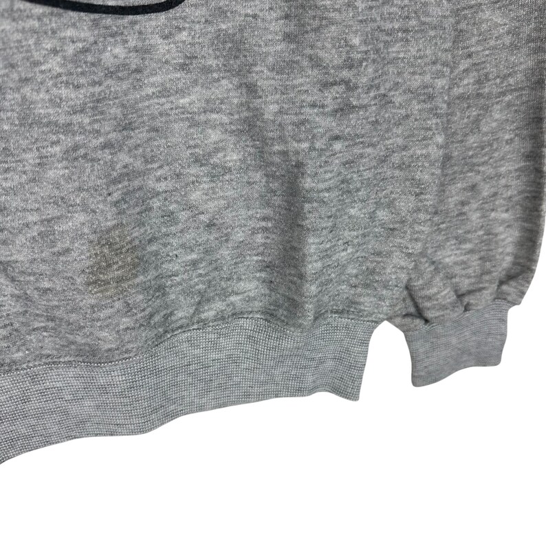 May include: A heather gray sweatshirt with a ribbed hem and cuffs. The fabric has a slightly textured appearance. There is a stain on the front of the sweatshirt. The bottom of the sweatshirt is visible.