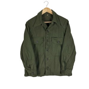 May include: An olive green long-sleeve shirt made of a thick, textured fabric. It features a pointed collar, a full-length front button closure, and two chest pockets with buttoned flaps. The sleeves have buttoned cuffs and reinforced shoulder seams.