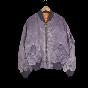 Puede incluir: Una chaqueta tipo bomber de color morado desteñido con cremallera metálica frontal, puños y dobladillo de canalé gris oscuro, dos bolsillos delanteros inclinados con cierre de corchete y un bolsillo utilitario en la manga izquierda.