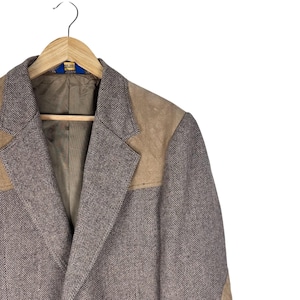 May include: A blazer featuring a brown and white herringbone pattern with tan suede shoulder panels and lapel accents. The garment has a notched lapel, a brown interior lining, and is displayed on a wooden hanger against a plain white background.