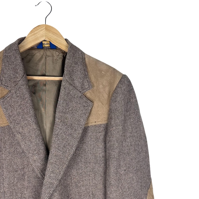 May include: A blazer featuring a brown and white herringbone pattern with tan suede shoulder panels and lapel accents. The garment has a notched lapel, a brown interior lining, and is displayed on a wooden hanger against a plain white background.