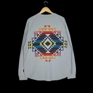 Op de afbeelding: Een lichtgrijs shirt met lange mouwen en een kleurrijk geometrisch ontwerp op de rug. Het ontwerp heeft een ruitvorm met driehoeken in rood, oranje, geel, groen, blauw en zwart. Het shirt hangt aan een houten hanger.