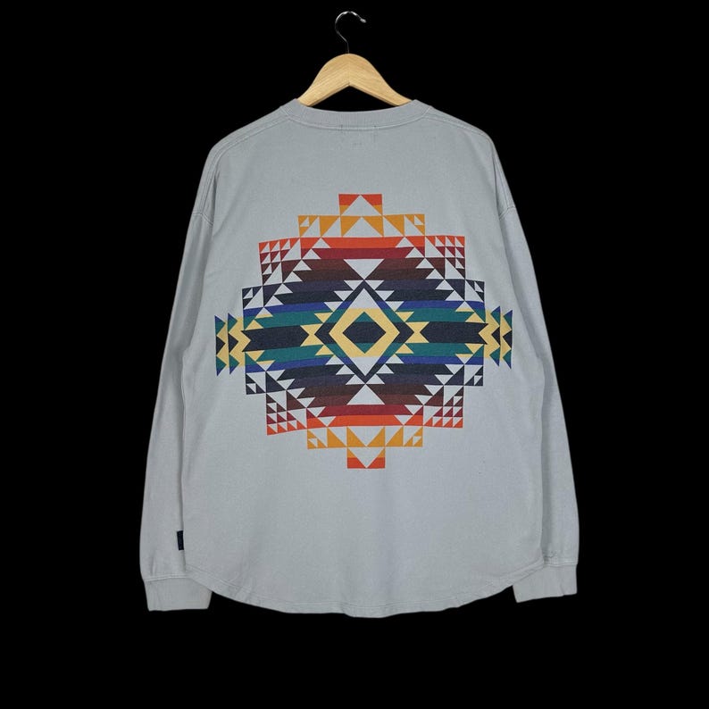 Puede incluir: Una camiseta de manga larga gris claro con un colorido dise&ntilde;o geom&eacute;trico en la espalda. El dise&ntilde;o presenta una forma de diamante con tri&aacute;ngulos en tonos rojos, naranjas, amarillos, verdes, azules y negros. La camiseta est&aacute; colgada en una percha de madera.