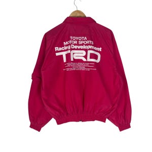 May include: A bright pink windbreaker jacket featuring a collar, long sleeves with elastic cuffs, and a zippered front. The back displays white printed text reading TOYOTA MOTOR SPORTS Racing Development TRD and additional descriptive text.