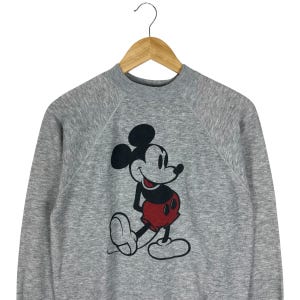 May include: A gray sweatshirt featuring a large graphic of Mickey Mouse. The cartoon character is depicted in black, red, and white. The sweatshirt has a crew neck and long sleeves, and is hanging on a wooden hanger.
