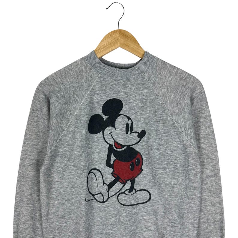 May include: A gray sweatshirt featuring a large graphic of Mickey Mouse. The cartoon character is depicted in black, red, and white. The sweatshirt has a crew neck and long sleeves, and is hanging on a wooden hanger.