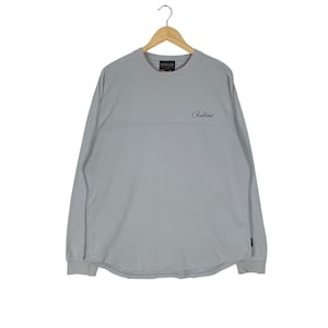 Puede incluir: Una camiseta de manga larga gris claro con cuello redondo, colgada de una percha de madera. La camisa tiene una costura horizontal en el pecho y la palabra "Pendleton" en cursiva. Una peque&ntilde;a etiqueta negra est&aacute; en la manga izquierda.