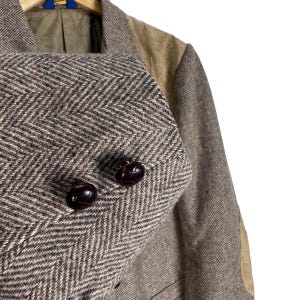 May include: A brown and tan blazer featuring a prominent herringbone pattern throughout the fabric. The garment includes contrasting tan suede-like shoulder panels and two dark brown, textured, round buttons on the front closure.