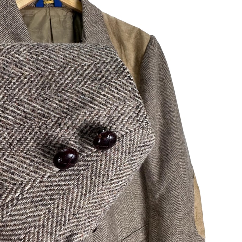 May include: A brown and tan blazer featuring a prominent herringbone pattern throughout the fabric. The garment includes contrasting tan suede-like shoulder panels and two dark brown, textured, round buttons on the front closure.