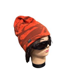 Op de afbeelding: Een oranje en bruin patroon beanie muts. De hoed heeft een donker rechthoekig label aan de zijkant. De hoed wordt gedragen door een mannequinhoofd met een zwarte zonnebril.