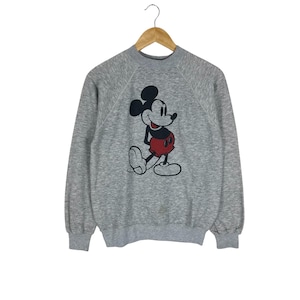 May include: A gray sweatshirt featuring a classic Mickey Mouse graphic. The cartoon character is depicted in black, red, and white. The long-sleeved top is hanging on a wooden hanger.