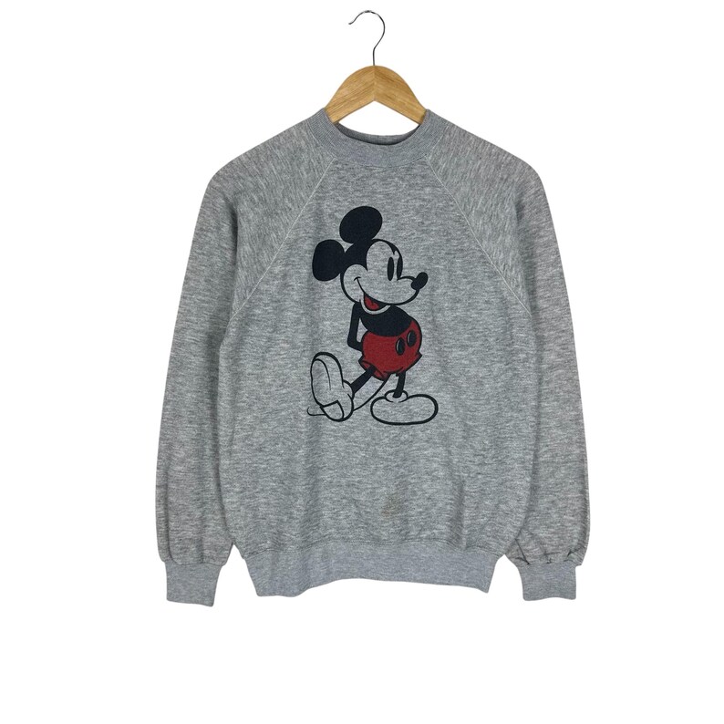 May include: A gray sweatshirt featuring a classic Mickey Mouse graphic. The cartoon character is depicted in black, red, and white. The long-sleeved top is hanging on a wooden hanger.
