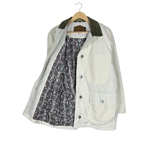 May include: Off-white jacket with a green corduroy collar and a paisley patterned lining. The jacket has two front pockets and is buttoned up. The jacket is hanging on a wooden hanger.