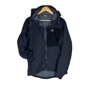 May include: A black hooded jacket with a full-length zipper and a small logo on the chest. The jacket has a two-tone design, with a black panel on the upper chest and shoulders. The jacket is hanging on a wooden hanger.