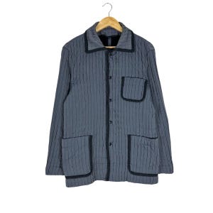 Puede incluir: Chaqueta acolchada gris con ribete negro y dos bolsillos delanteros. La chaqueta tiene una botonadura frontal y un cuello. La chaqueta está colgada en una percha de madera.