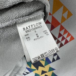 Puede incluir: Primer plano de una etiqueta de sudadera gris con el texto "BAYFLOW BM2108MN03". La etiqueta incluye informaci&oacute;n sobre la talla y la composici&oacute;n de la tela. El fondo presenta un patr&oacute;n geom&eacute;trico en naranja, amarillo y blanco.