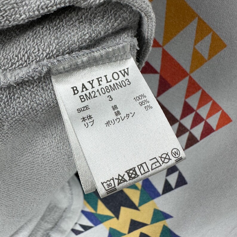 Puede incluir: Primer plano de una etiqueta de sudadera gris con el texto "BAYFLOW BM2108MN03". La etiqueta incluye informaci&oacute;n sobre la talla y la composici&oacute;n de la tela. El fondo presenta un patr&oacute;n geom&eacute;trico en naranja, amarillo y blanco.