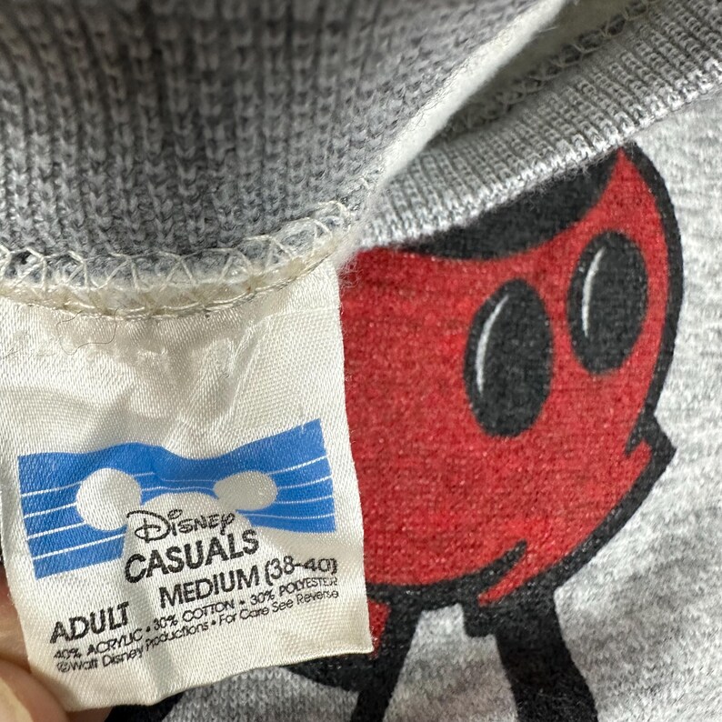 May include: Close-up of a gray Disney Casuals sweatshirt with a red Mickey Mouse graphic. The tag reads "ADULT MEDIUM (38-40)" and lists the fabric content as 40% acrylic, 30% cotton, and 30% polyester.