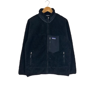 May include: Black fleece jacket with a full zipper closure, a stand-up collar, and a chest pocket. The jacket has two side pockets with zippers and a small logo patch on the chest pocket. The jacket is hanging on a wooden hanger.