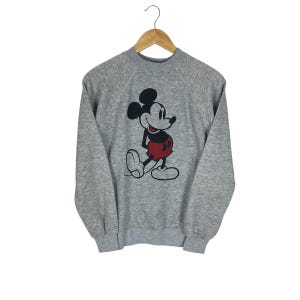 May include: A gray sweatshirt featuring a classic Mickey Mouse graphic. The cartoon character is depicted in black, red, and white. The sweatshirt is hanging on a wooden hanger against a white background.