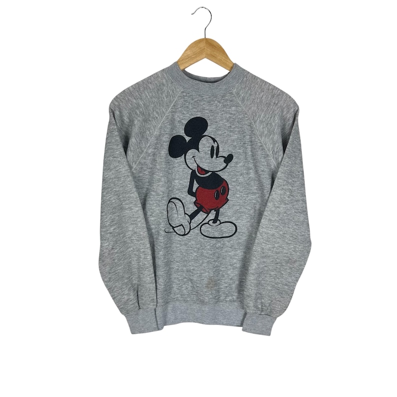 May include: A gray sweatshirt featuring a classic Mickey Mouse graphic. The cartoon character is depicted in black, red, and white. The sweatshirt is hanging on a wooden hanger against a white background.