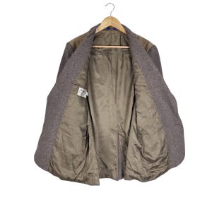 May include: A brown tweed blazer with a textured herringbone pattern, featuring a smooth tan interior lining, a single interior chest pocket, and a white brand label stitched inside the left panel, all displayed on a wooden clothes hanger.