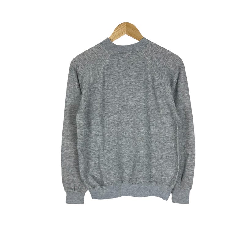 May include: A heather gray sweatshirt with long sleeves and a crew neck, hanging on a wooden hanger. The sweatshirt has a ribbed hem and cuffs, and white stitching along the shoulders and sleeves.