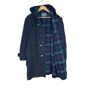 May include: A dark navy blue duffle coat with a hood, featuring four wooden toggle closures and a green, blue, and red plaid interior lining. The coat includes a label at the neck that reads Gloverall, Made in England.