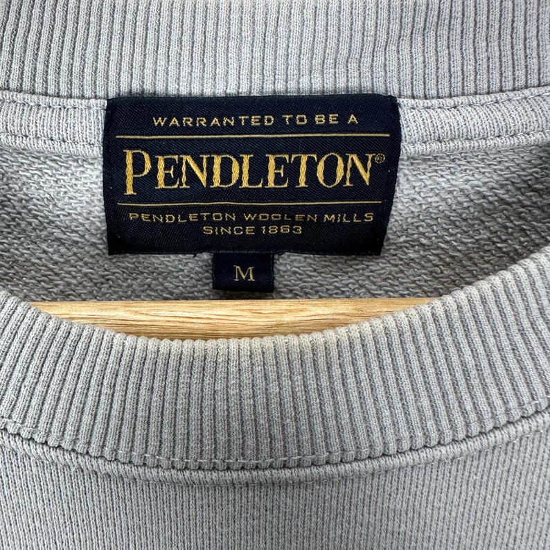 Puede incluir: Primer plano de una sudadera gris con cuello redondo acanalado. La etiqueta negra dice "WARRANTED TO BE A PENDLETON PENDLETON WOOLEN MILLS SINCE 1863" en letras doradas. La talla "M" est&aacute; impresa debajo.